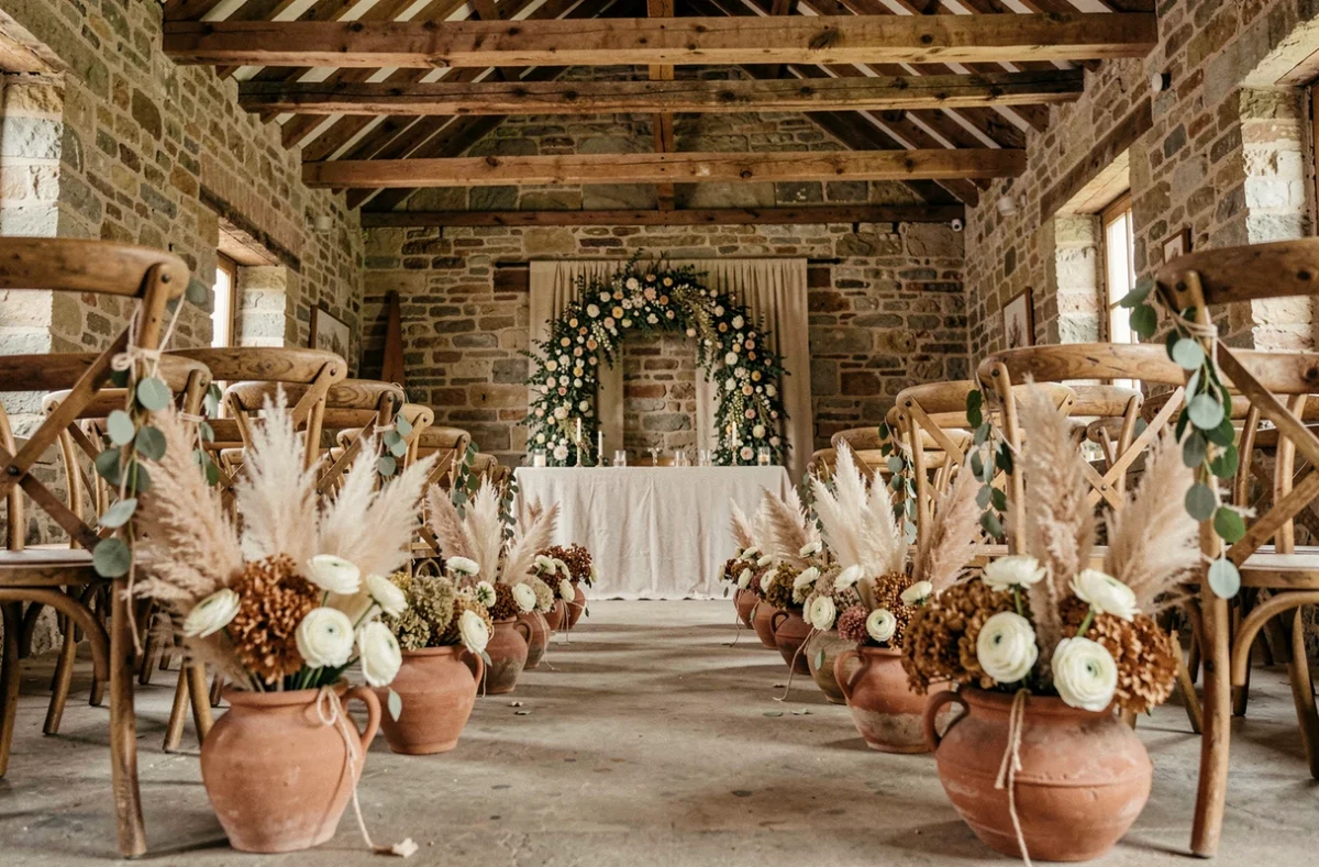 Allée de cérémonie de mariage rustique en grange de pierre avec herbes de la pampa, fleurs séchées et chaises en bois